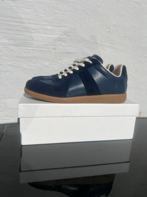 Marinblå Maison margielas - Snygga marinblå sneakers i läder med vita skosnören och klassisk gummisula. Skorna har en låg profil och stilrena detaljer, perfekt för dig som gillar en clean look. Insidan är ljusbeige och designen är tidlös.