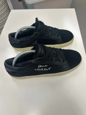 Svarta Saint Laurent sneakers - Snygga svarta sneakers från Saint Laurent med vit logga på sidan. Skorna har lågt skaft, svarta skosnören och vit sula. Tillverkade i canva och läder för en stilren och trendig look.