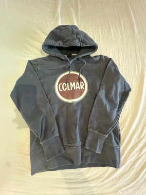 Blå Colmar hoodie med logga - Snygg blå hoodie från Colmar med stor rund logga i vitt och vinrött på bröstet. Klassisk modell med huva och dragsnören samt ribbade muddar vid ärmar och nederkant. Perfekt nu när det börjar bli varmare ute. I storlek S. Passar ungefär 158-172cm
