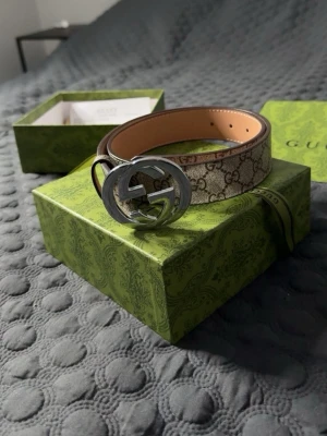 GG Supreme bälte med silverdetalj - Snyggt GG Supreme bälte i beige och brunt med klassiskt monogrammönster och stor silverfärgad GG-logga som spänne. Bältet är gjort i skinn och har exklusiv känsla, perfekt för att lyfta vilken outfit som helst. Skärpet helt nytt och allt medföljer, låda och dustbag. Skriv vid frågor!