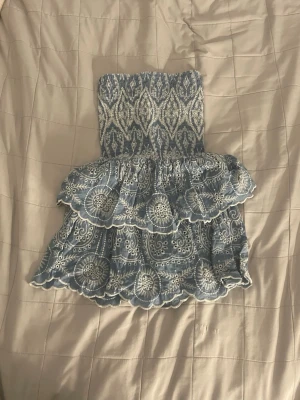 Blåvit axelbandslös kortklänning - Supersöt blåvit axelbandslös kortklänning med volangkjol och broderade detaljer. Klänningen har ett vackert mönster och smockad överdel som ger en bekväm passform. Perfekt för varma dagar och semester. 