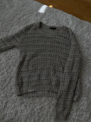 Grå stickad tröja med hålmönster - Säljer en grå stickad tröja med snyggt hålmönster och rund hals. Tröjan har ribbade muddar vid ärmslut och nederkant, vilket ger en skön passform. Perfekt för lager på lager och enkel att styla till olika outfits.