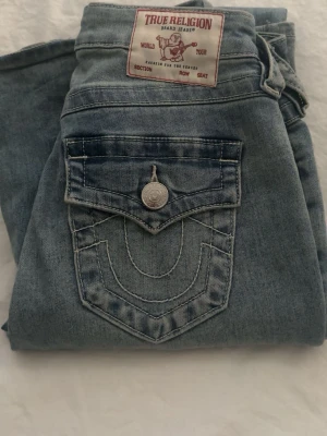 True Religion ljusblå bootcut jeans - Säljer ett par ljusblå bootcut jeans från True Religion med ikonisk fickdetalj och kontrastsömmar. Jeansen har en klassisk femficksmodell, knappstängning och ett snyggt tvättat utseende. Perfekta för dig som gillar en avslappnad men trendig stil. 