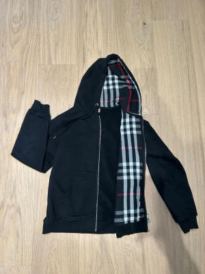 Svart hoodie med dragkedja från Burberry - Säljer en svart hoodie från Burberry med dragkedja och klassiskt rutigt foder i huvan. Hoodien har fickor framtill och justerbar huva med snören. Perfekt för dig som gillar stilrena plagg.