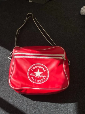 Röd Converse Chuck Taylor - Röd axelväska från Converse i klassisk retrostil med vit piping och stor logga på framsidan. Väskan har justerbar axelrem, ett stort huvudfack och en dragkedjeförsedd ytterficka. Materialet är glansigt konstläder som ger en sportig vibe.