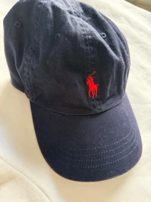 Marinblå Ralph lauren keps röd logo - Marinblå keps från ralph lauren med röd logo, använd 1 sommar men har väl tagit hand om den så den är fräsch. Frakten står köparen för :)