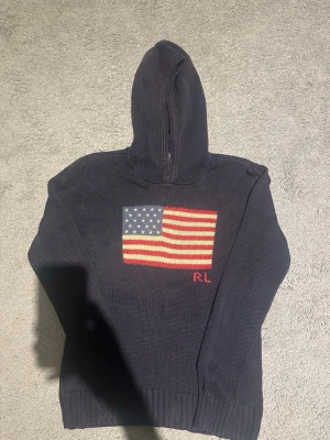 Ralph Lauren stickad hoodie - Snygg mörkblå hoodie från Ralph Lauren med stickad amerikansk flagga på bröstet och RL-broderi i rött. Tillverkad i tjockt stickat material som ger en exklusiv känsla. Perfekt för dig som vill ha en klassisk men ändå statement look. Storlek 12-16 år men passar dig som är upp till 175 cm. Bra skick! Priset är ej hugget i sten!!