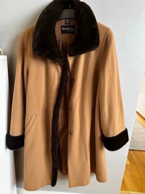 Kamelbrun kappa med fuskpäls Berghaus - Snygg kamelbrun kappa från Berghaus med bred krage och muddar i mörkbrun fuskpäls. Kappan har knäppning framtill, två sidofickor och ett mjukt innerfoder. Perfekt för kalla dagar och ger en klassisk vintagekänsla. Rök och djurfritt hem. Pruta gärna!🌸