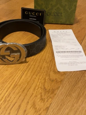 Svart Gucci bälte  - Säljer ett svart skärp från Gucci med det klassiska GG-spännet i silver. Skärpet har diskret mönster med GG-loggor längs hela bandet och är gjort i skinn. Perfekt accessoar för att lyfta vilken outfit som helst. Den är 110 cm 