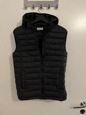 Svart dunväst med huva från Mango - Snygg svart dunväst från MNG med huva och dragkedja framtill. Västen har två fickor på sidorna och quiltad design som ger en modern look. Perfekt att slänga över en hoodie eller tröja för extra värme. Materialet är lätt och smidigt.