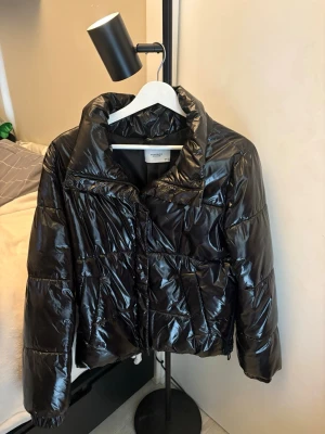 Svart pufferjacka från Reserved - Snygg svart pufferjacka från Reserved Eco Aware med glansig finish och hög krage. Jackan har dragkedja framtill, två fickor och en boxig, trendig passform. Perfekt för kalla vinterdagar och ger en cool look till din outfit.