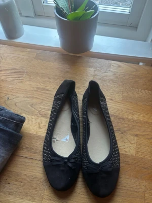 Svarta ballerinaskor med strassdetaljer - Säljer ett par svarta ballerinaskor med små glittrande strassdetaljer längs sidorna och en söt rosett framtill. Skorna har rundad tå och är i ett mjukt material som ser ut som mocka. Perfekta för dig som vill ha något stilrent men ändå lite extra.