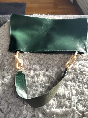 Grön kuvertväska med kedjedetalj - Stilren grön kuvertväska i glansigt konstläder med rektangulär form. Väskan har ett brett axelband i matchande grönt och coola kedjedetaljer i beige plast. Perfekt accessoar för att lyfta din outfit. Knappt använd. 37x22 cm