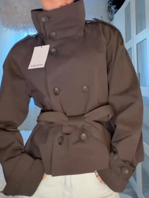 Kort brun trenchcoat från Bershka - Snygg kort trenchcoat i mörkbrunt från Bershka med dubbelknäppning, hög krage och knytskärp i midjan. Jackan har axelklaffar och dekorativa knappar vid ärmsluten. Perfekt för dig som vill ha en trendig och stilren look. Helt oanvänd med prislapp i storlek S