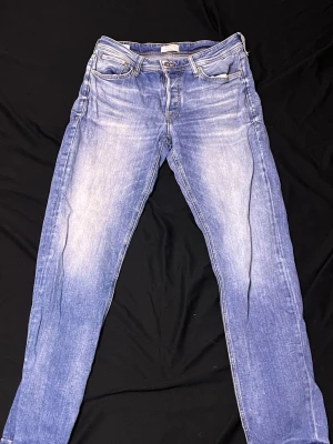 Blå Tapered jeans Mike 32/34 - Säljer ett par blå tvättade jeans från Mike i modellen Tapered. Jeansen har klassisk femficksdesign, dragkedjegylf och snygga slitningar framtill. Perfekta för dig som gillar en avslappnad men ändå stilren look.