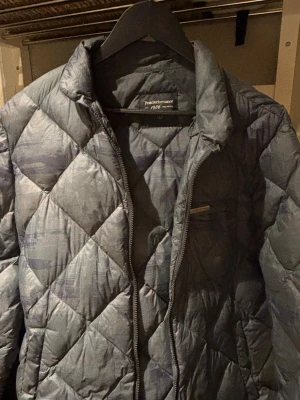 Grå dunjacka Peak Performance L - Kamouflage  - Snygg grå dunjacka från Peak Performance med diskret mönster och quiltad design. Jackan har dragkedja framtill, hög krage och en bröstficka med dragkedja. Perfekt för kalla höst/vår/vinterdagar och riktigt skön att bära.