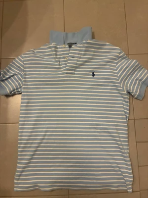 Blåvit randig piké från Ralph Lauren - Snygg blåvit randig pikétröja från Ralph Lauren med klassisk krage och knappar framtill. Tröjan har det ikoniska Polo-logotypen broderad på bröstet och är tillverkad i mjuk bomull. Perfekt för en avslappnad men stilren look.