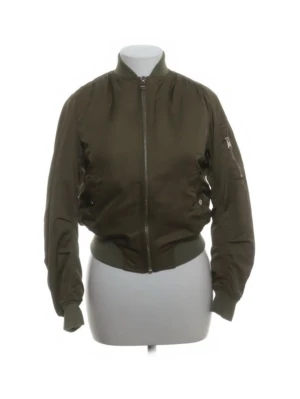 Grön bomberjacka med dragkedja - Snygg olivgrön bomberjacka med klassisk ribbad mudd i midja och ärmslut. Jackan har en rak passform, silverfärgad dragkedja framtill och fickor på sidorna samt en ficka med dragkedja på ena ärmen. Tillverkad i ett glansigt material som ger en cool look.
