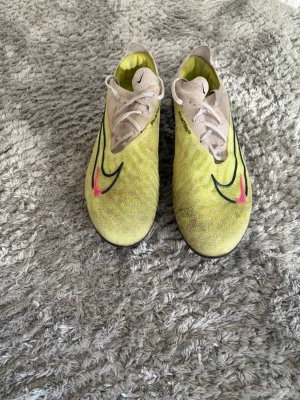 Nike Phantom GT2 gula fotbollsskor - Säljer ett par Nike Phantom GT2 fotbollsskor i gul och beige med svarta och rosa detaljer. Skorna har snörning, låg profil och ovandel i syntetmaterial för bra bollkontroll. Perfekta för dig som vill sticka ut på planen.