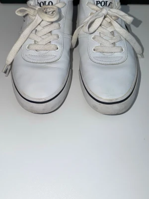 Vita Polo Ralph Lauren sneakers - Snygga vita sneakers från Polo Ralph Lauren med klassisk låg profil och vita snören. Skorna har diskret logga på sidan och märkesdetaljer på plösen. Tillverkade i slätt läder med gummisula och svart rand runt sulan för en clean look.