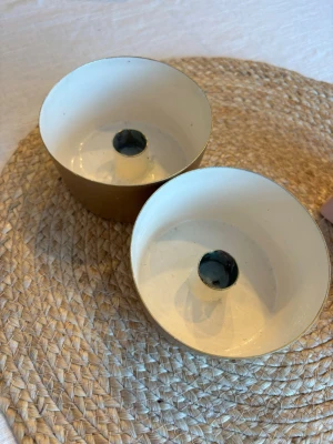 Guldiga metall-ljushållare, 2-pack - Två stilrena ljushållare i metall med guldig utsida och vit insida. Runda i formen och perfekta för värmeljus. Passar fint som dekoration på bord eller fönsterbräda. Säljes som ett set om två stycken.