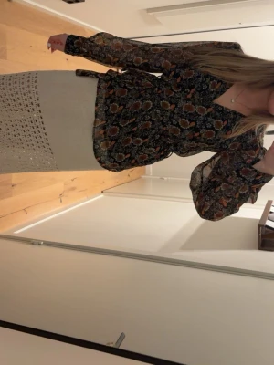 Blommig blus med knytband - Säljer en blommig blus i tunnt, transparent material med svart botten och mönster i orange, vitt och grönt. Blusen har långa ärmar och ett knytband i midjan för en snygg siluett. Perfekt för dig som gillar boho-stil och vill ha något unikt i garderoben.