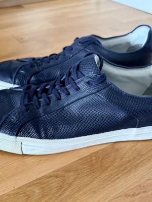 Mörkblå sneakers med reptilmönster - Zara - Snygga mörkblå sneakers från Zara med reptilmönstrad yta och vita sulor. Skorna har klassisk låg profil, matchande mörkblå snören och en stilren look som passar till det mesta. Materialet ser ut att vara konstläder eller läder.