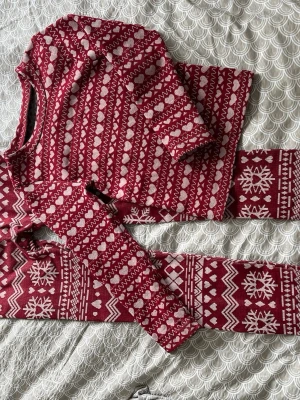 Vinröd pyjamas med vintermönster - Mysig tvådelad pyjamas i vinrött med vita hjärtan, snöflingor och zigzag-mönster. Tröjan har långa ärmar och rund hals, byxorna är långa och matchar med samma vinterinspirerade print. Perfekt för kalla kvällar och julmys. Kan användas som underställ också. 
