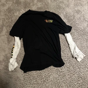 Svart Vans topp med vita ärmar - Cool svart topp från Vans med vita långa ärmar i lager-på-lager stil. Liten Vans-logga i gult och rött på bröstet. Ärmarna har tryck i gult och svart. Perfekt för en avslappnad streetwear-look.