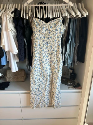 Blommig långklänning med snörning - Söt långklänning med smala axelband och blått blommönster på vit botten. Klänningen har en feminin v-ringning och snörningsdetalj i ryggen som ger en snygg passform. Perfekt för sommarens alla tillfällen.