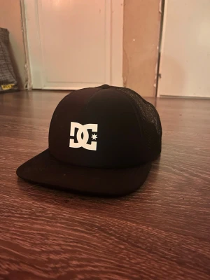 Svart DC keps med mesh - Säljer en svart keps från DC med vit logga framtill. Kepsen har platt skärm och mesh på sidorna och bak för extra ventilation. Justerbar baktill för perfekt passform. Perfekt för dig som gillar streetwear och skate.