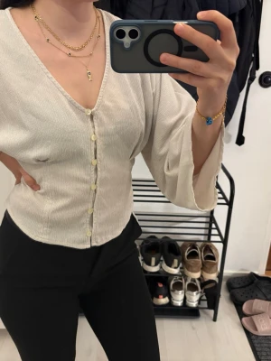  blus med vida ärmar från Lindex - Säljer en krämvit blus från Lindex med v-ringning och knappar framtill. Blusen har långärmade, vida ärmar och är gjord i ett lätt, strukturerat tyg som ger en avslappnad men chic look. Perfekt att styla med både jeans och kjol.