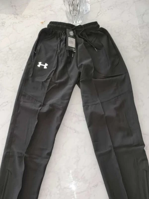 Svarta mjukisbyxor från Under Armour - Säljer ett par svarta mjukisbyxor från Under Armour med dragsko i midjan och resår. Byxorna har sidofickor och en vit logga på vänster ben. Perfekta för chill eller träning, tillverkade i ett mjukt och bekvämt material.
