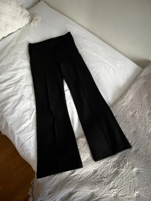 Svarta utsvängda kostymbyxor - Säljer ett par svarta kostymbyxor med utsvängda ben och pressveck. Byxorna har hällor för skärp och en klassisk midja. Perfekta för en stilren och trendig look.