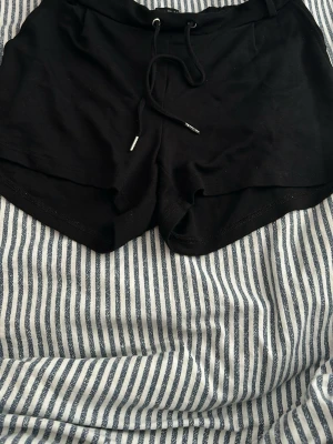 Svarta shorts med snörning - Svarta shorts i mjukt material med snörning i midjan och elastisk linning. Perfekta för sommardagar eller chill hemma. Klassisk passform och enkel design som funkar till det mesta.