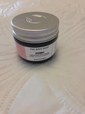 Vitamin E dagkräm från The Body Shop - Fuktgivande dagkräm med vitamin E från The Body Shop. Kommer i en rund burk med silverfärgat lock och etikett i vitt och rosa. Innehåller 50 ml och är perfekt för att återfukta huden under dagen. Endast öppnad och aldrig använd.