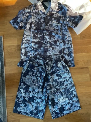 Blommigt set från Louis Vuitton - Säljer ett snyggt set från Louis Vuitton med kortärmad skjorta och matchande shorts. Setet har ett blått och vitt blommönster och Louis Vuitton-logga broderad på bröstet och shortsen. Materialet ser ut som denim eller bomullsmix. Perfekt för dig som vill sticka ut!