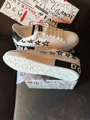 Dolce & Gabbana vita sneakers - Snygga vita sneakers från Dolce & Gabbana med svarta stjärnor och textdetaljer på sidorna. Skorna har svarta hälpartier och vita snören. Sulan är präglad med märkets logga och mönster. Tillverkade i läder för en exklusiv känsla.