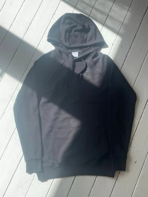Svart hoodie från Gymshark - Snygg svart hoodie från Gymshark med broderad logga på bröstet och känguruficka framtill. Hoodien har dragsko i huvan och ribbade muddar vid ärmslut och nederkant. Perfekt för en sportig och avslappnad stil.