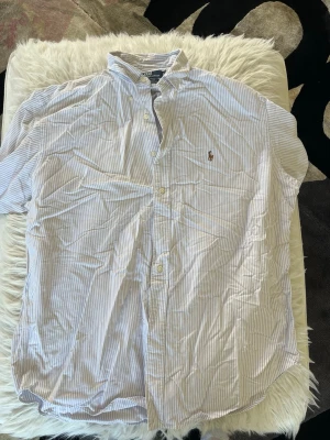  Regnbåg randig skjorta från Polo Ralph Lauren - Klassisk långärmad skjorta från Polo Ralph Lauren i vitt med tunna grå ränder. Skjortan har button-down krage, broderad logga på bröstet och är tillverkad. Perfekt för en clean och stilren look.