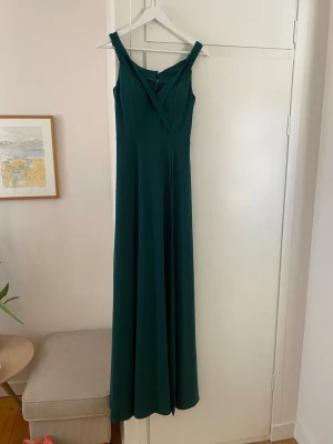 Mörkgrön balklänning med v-ringning - Mörkgrön balklänning med breda axelband och snygg v-ringning framtill. Klänningen har en figurnära överdel och ett långt, flödande kjolparti med en slit på vänster ben. Skriv för fler bilder/bilder på.