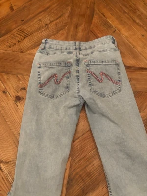Ljusblå bootcut jeans med slitningar - Säljer ett par ljusblå bootcut jeans med slitna detaljer på knäna och broderade bakfickor med rosa zigzag-mönster. Jeansen har klassisk femficksdesign och är tillverkade i denim. Perfekta för en avslappnad och trendig look. Dem är lite fuktiga på bilden som gör att dom ser smutsiga ut. Knappt använda.