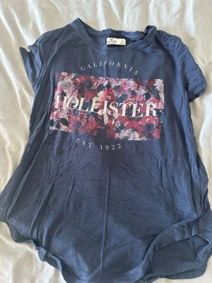 Blommig blå t-shirt från Hollister - Snygg blå t-shirt från Hollister med blommigt tryck och texten 'California' och 'Est 1922' på bröstet. T-shirten har rund hals och korta ärmar, perfekt för en avslappnad look. Tillverkad i mjuk bomull för skön känsla hela dagen.