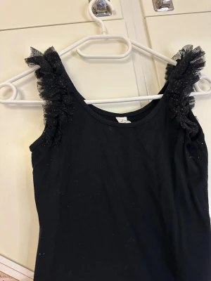 Svart linnetopp med volangaxlar - Snygg svart linnetopp med ribbad struktur och volangdetaljer i mesh på axlarna. Toppen har ärmlös design och en enkel, tight passform som ger en trendig look. Perfekt att styla med jeans eller kjol för en cool outfit.