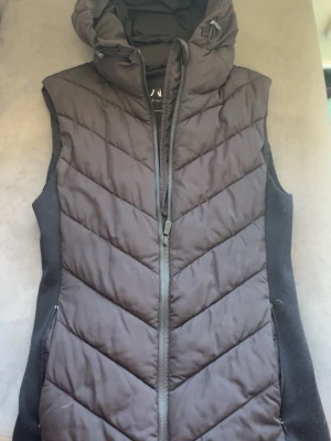 Svart dunväst med huva från zara  - Snygg svart dunväst från Zara  med dragkedja framtill och stor, mysig huva. Västen har ett quiltat mönster i chevron-stil och sidopaneler i stretchigt tyg för extra komfort. Perfekt för lager-på-lager och streetwear-vibe.