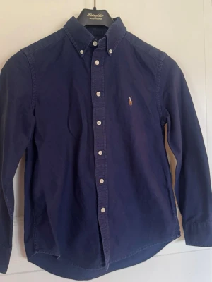 Marinblå skjorta från Ralph Lauren - Klassisk marinblå skjorta från Ralph Lauren med button-down krage och vita knappar. Skjortan har lång ärm och den ikoniska broderade loggan på bröstet. Tillverkad i mjuk bomull för en bekväm känsla och snygg passform.