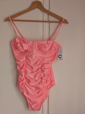 Cubus Baddräkt i ett stycke Storlek M - Snygg rosa baddräkt från Cubus med tunna axelband och rynkade detaljer framtill. Baddräkten har bygel under bysten för extra stöd och är tillverkad i ett glansigt stretchmaterial som ger en lyxig känsla. Perfekt för strand eller poolhäng.