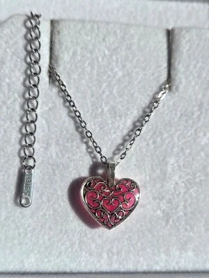 Halsband med hjärthänge som lyser lila i mörker - Snyggt halsband i silverfärg med ett hjärtformat hänge i rosa och dekorativa snirkliga detaljer. Hänget lyser i lila när det är mörkt efter att ha varit i dagsljus, direkt solljus ger absolut starkast effekt efter bara en kort stund men även vanligt ljus utifrån fungerar också vilket betyder att effekten sköter sig själv när du bär smycket. Kedjan är i rostfritt stål. Perfekt accessoar för att sticka ut.