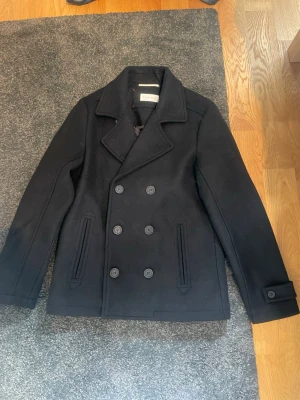 Marinblå skepparkavaj från Filippa K - Stilren marinblå skepparkavaj från Filippa K med dubbelknäppning och bred krage. Klassisk modell med två framfickor och dekorativa knappar vid ärmsluten. Tillverkad i ullblandning som håller värmen under kyliga dagar.