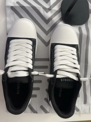 Svarta och vita sneakers från NUMERIS - Snygga sneakers från NUMERIS i svart och vitt med vita snören och kontrastsömmar. Ovandelen är i slätt och texturerat syntetmaterial, och sulan är vit med räfflad kant. Klassisk låg modell som passar till många stilar. Storlek 42 och har enbart använts vid få tillfällen, kom gärna med prisförslag 